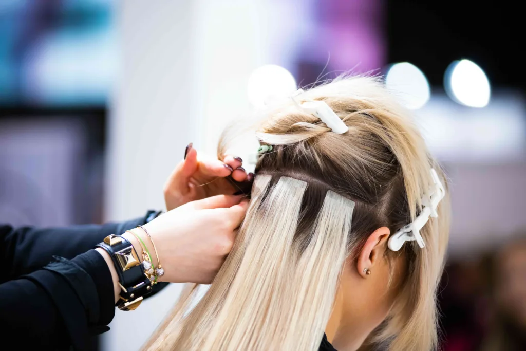 A young woman getting custom volume hair extensions at Panache Hair Salon in Meridian, ID. View of the back of her head of brown hair, receiving long, blonde hand tied and beaded wefts of real human hair. This is a great treatment for thinning hair loss, restoring confidence and beauty. 