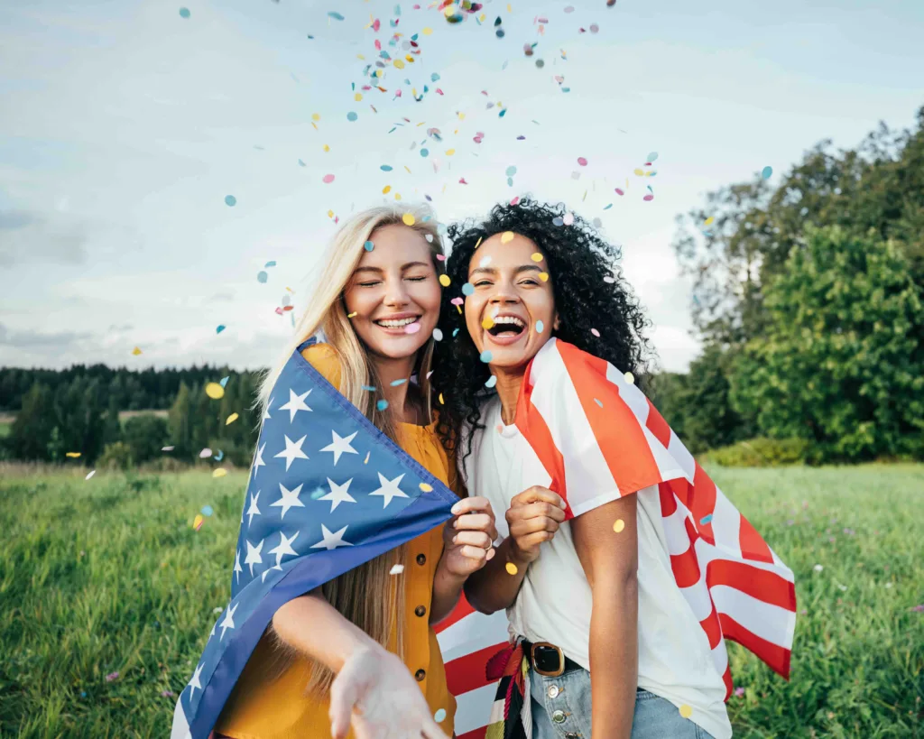 2 happy, young women draped in an American flag, celebrating the 4th of July and America 250 by reading the definitive history of the United States of America, as told through our nation's most popular hairstyles throughout our storied 250 year history.