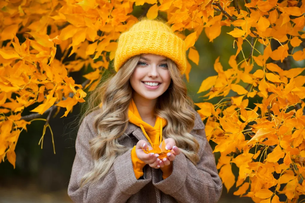 A young woman standing outside during Fall, holding orange leaves, in a yellow hat, smiling. Showing the benefits of an updated Autumn skincare routine from Panache Hair Studio & Day Spa in Meridian, Idaho.
